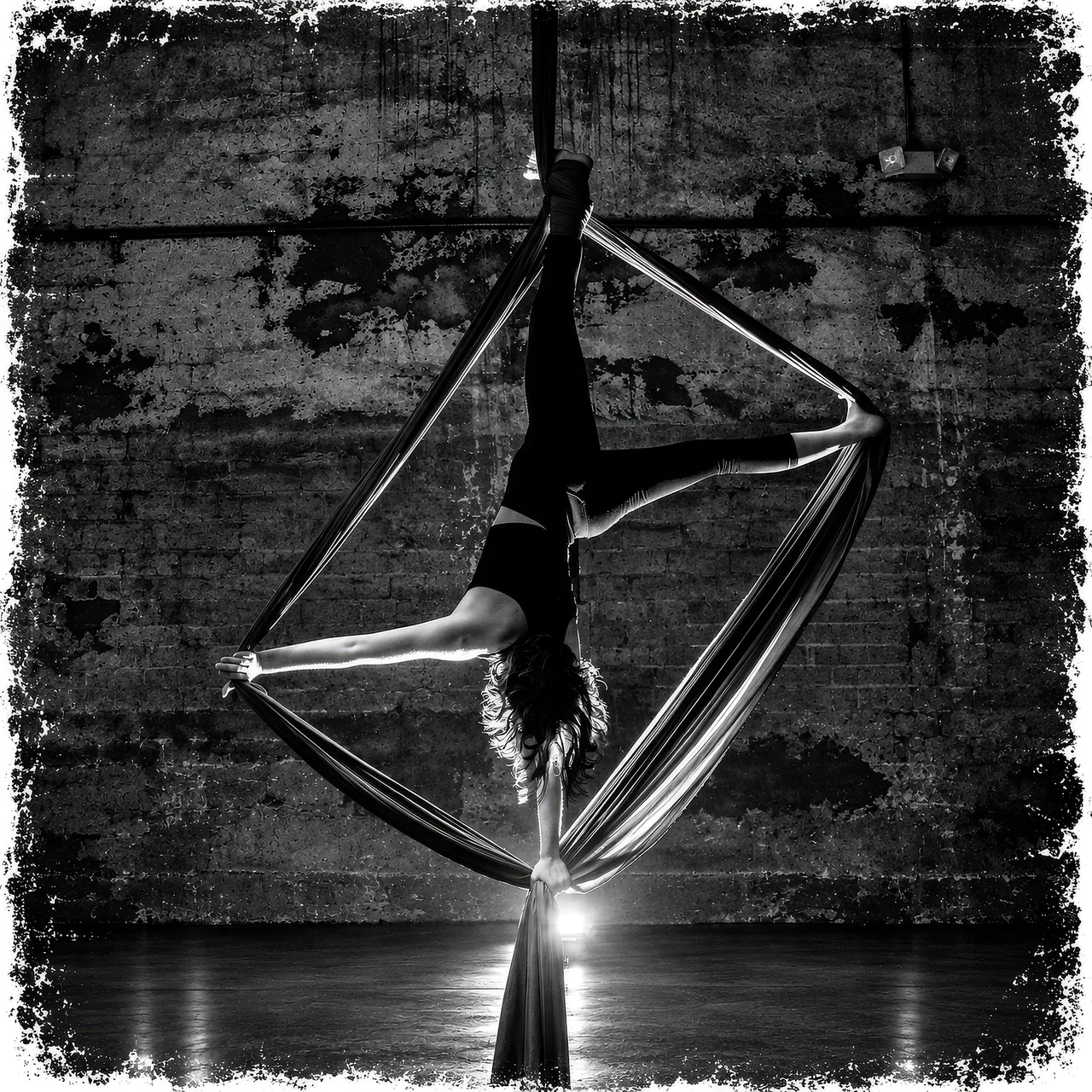 Black-and-white photograph of an aerial performer suspended in fabric, framed with a textured border.